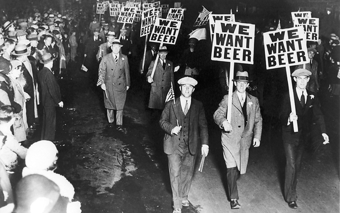 A large crowd of protesters marching through city streets at night, carrying signs reading 'We Want Beer' during Prohibition in the early 1930s.